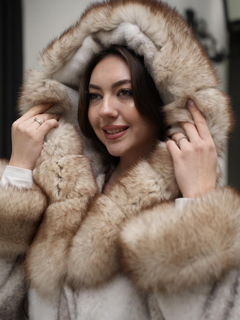 May include: A hooded fur coat with a light brown and cream color scheme. The coat features a large hood and trim around the collar and cuffs. The fur appears soft and luxurious, creating a warm and stylish outerwear piece.