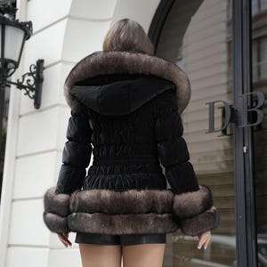 以下が含まれることがあります： 黒いパファージャケット、毛皮トリムのフードと袖口付き。 ジャケットはウエストが絞られており、キルティングデザインです。 モデルは黒いショートパンツを着用しています。 スタイリッシュなアウターウェアです。