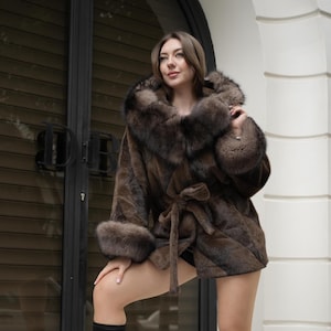 May include: A brown fur coat with a hood and tie closure. The coat has fur trim on the hood and cuffs. The model is wearing the coat outdoors.