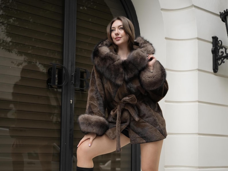May include: A brown fur coat with a hood and tie closure. The coat has fur trim on the hood and cuffs. The model is wearing the coat outdoors.