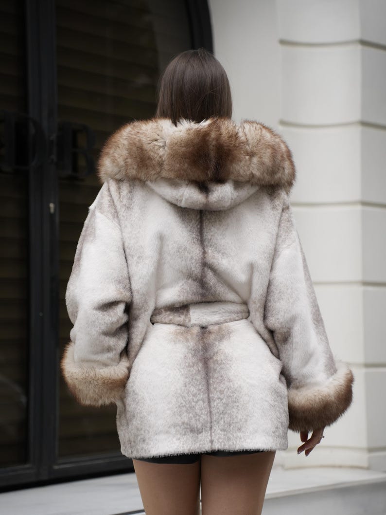 May include: A hooded, faux fur coat in shades of white and brown, featuring a belted waist and fur trim on the hood and cuffs. The coat is designed for warmth and style, offering a luxurious look.