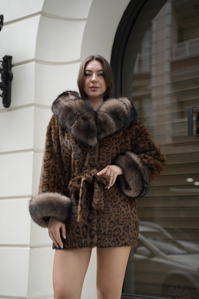 May include: A brown and black leopard print coat with a fur collar and cuffs. The coat is belted at the waist and has a luxurious appearance. The model is wearing the coat in an outdoor setting.