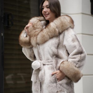 May include: A light-colored faux fur coat with a hood and cuffs trimmed with brown fur. The coat has a matching belt and a pocket. The coat is knee-length and appears to be made of a soft, plush material.