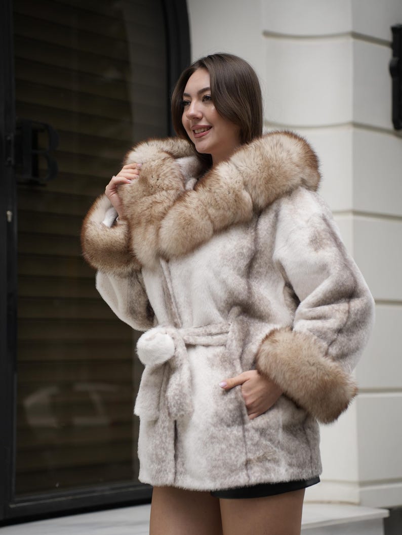 May include: A light-colored faux fur coat with a hood and cuffs trimmed with brown fur. The coat has a matching belt and a pocket. The coat is knee-length and appears to be made of a soft, plush material.