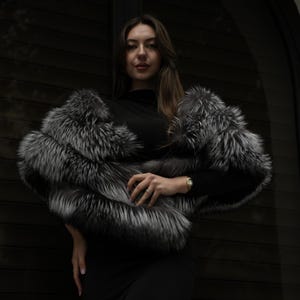 May include: A luxurious silver and black fur wrap, worn over a black top. The wrap features a fluffy texture and a gradient of silver and black hues. The backdrop is a dark, textured surface.