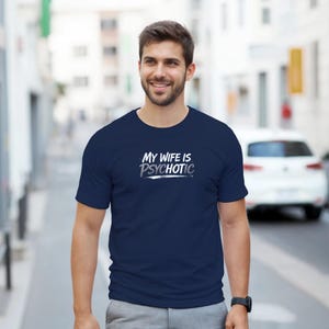 May include: Navy blue t-shirt with the text "MY WIFE IS PSYCHOTIC" in white and light blue. The shirt is short-sleeved and worn by a person in an outdoor setting.