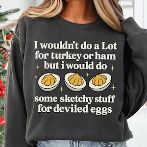 Op de afbeelding: Donkergrijze sweatshirt met de tekst "I wouldn't do a Lot for turkey or ham but I would do some sketchy stuff for deviled eggs." Het ontwerp bevat illustraties van gevulde eieren en sterren.