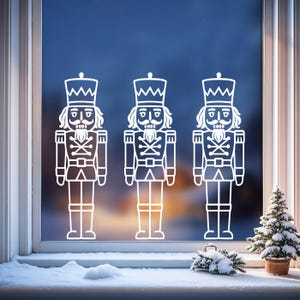 Puede incluir: Tres imágenes blancas de cascanueces se muestran en una ventana. Los cascanueces están en fila, cada uno con un uniforme detallado y un sombrero alto. Un pequeño árbol de Navidad y nieve son visibles en el alféizar de la ventana.