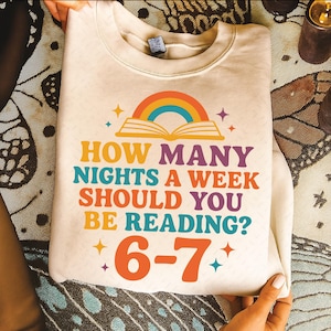 Op de afbeelding: Crèmekleurige sweatshirt met de tekst "HOW MANY NIGHTS A WEEK SHOULD YOU BE READING?" in oranje, teal en paars. Een regenboog en open boek grafiek staat boven de tekst, met de cijfers "6-7" in oranje eronder.