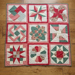 Puede incluir: Una colcha de patchwork cuadrada con nueve bloques distintos dispuestos en una cuadrícula. La colcha muestra una variedad de patrones geométricos y florales en tonos rojos, verdes y crema, con cada bloque separado por tiras rojas.