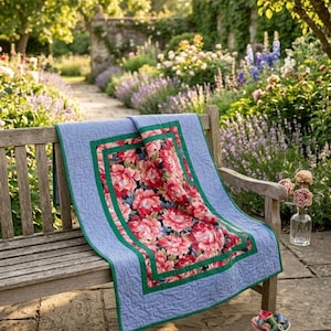 May include: A rectangular quilt featuring a central panel of dark fabric with a vibrant red and pink floral pattern, framed by a solid green border and a wider light blue outer border with a quilted texture draped over a wooden bench.