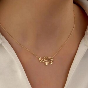 May include: Gold-coloured necklace with an elephant pendant, inscribed with the name "Kinsley". The necklace is worn against a white shirt.