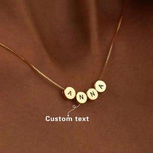May include: A gold-coloured necklace with a fine chain. The necklace displays five circular pendants, each with a letter spelling "ANNA". The words "Custom text" are written below the pendants.
