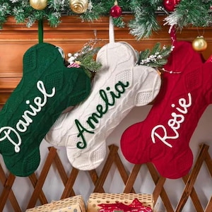 May include: Three Christmas stockings shaped like dog bones hang from a decorated mantel. The stockings are green, white, and red, with the names "Daniel", "Amelia", and "Rosie" embroidered in white script. Festive holiday decor surrounds the stockings.