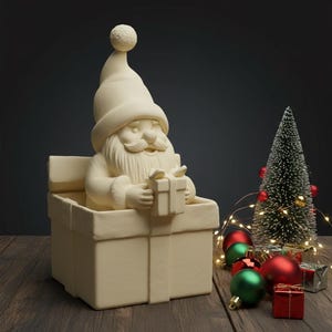 Könnte beinhalten: Eine cremefarbene Weihnachtsmannfigur, die aus einer Geschenkbox auftaucht und ein kleines Geschenk hält. Ein Miniatur-Weihnachtsbaum, rote und grüne Kugeln sowie Lichterketten sind auf einer Holzoberfläche angeordnet und schaffen eine festliche Urlaubsszene.