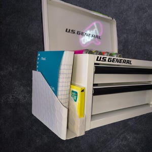 May include: A white U.S. General metal tool chest with two drawers and an open lid. Attached to the side is a white plastic organizer holding a teal Mead notebook and a yellow Pen+Gear notepad. The U.S. General logo is printed on the lid and front.