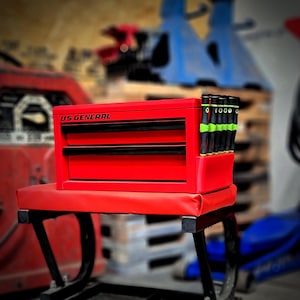 May include: A red US GENERAL tool chest with black drawer handles and a set of black and green screwdrivers. The tool chest is on a red padded stool with black legs. The background is blurred.