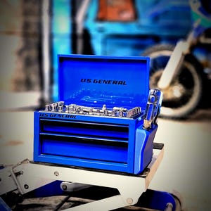 May include: A bright blue US GENERAL toolbox with open top and drawers, filled with various tools. The toolbox is on a white and blue lift, with the word PITTSBURGH visible. The background includes a blurred blue vehicle and motorcycle.