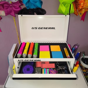 May include: A white U.S. General metal tool box used as a desk organizer. The open lid and drawer reveal compartments filled with neon highlighters, colorful sticky notes, yellow pencils, a purple tape dispenser, red erasers, and metal binder clips.