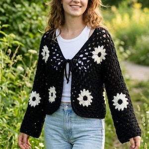 May include: Black crochet cardigan with white daisy flower accents. The open-front sweater has long sleeves and a tie closure. The model is wearing a white top and light blue jeans. The background is a green field.