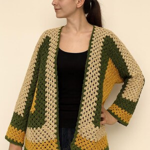 May include: A handmade crochet cardigan in a granny square pattern. The open-front sweater features a color block design with olive green, mustard yellow, and beige panels. The long-sleeved garment is a cozy, bohemian style.
