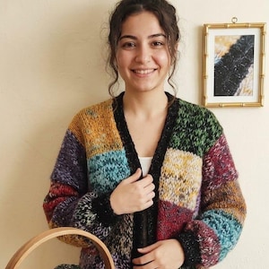 Może przedstawiać: Wielokolorowy kardigan w stylu patchwork z czarnym wykończeniem. Posiada kwadraty w kolorach żółtym, zielonym, niebieskim i czerwonym. Drewniany kosz zawiera kulki wełny w różnych kolorach. Widoczne są również druty do robienia na drutach.