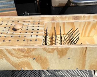 Puede incluir: Una caja de madera con una tapa con bisagras y un compartimento con clavos que sobresalen. La tapa tiene un pomo en la parte superior y una cuadrícula de clavos.