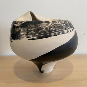 Peut inclure: Un vase en céramique au design abstrait unique. Il présente un corps arrondi, un sommet évasé et un pied en forme de piédestal. La partie supérieure est crème, tandis que la moitié inférieure est noire, avec des coups de pinceau noirs audacieux sur la section crème.