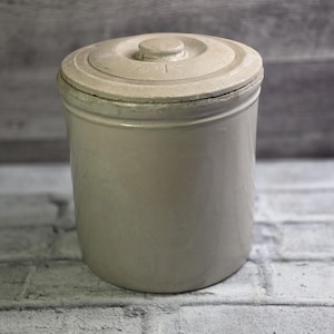 May include: A cream-coloured stoneware crock with a matching lid. The cylindrical container has a slightly tapered shape and a lid with a central knob. The crock appears to be in good condition, suggesting it is well-preserved.