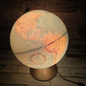 Peut inclure: Une lampe globe vintage éclairée, avec une base dorée. Le globe affiche une carte détaillée avec les pays et les continents, éclairée de l'intérieur. La lampe est branchée et prête à l'emploi, idéale pour un bureau.