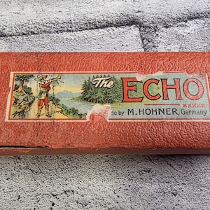 May include: A vintage red rectangular box with a label featuring a man playing a horn near a lake and the text "The ECHO" and "made by M. Hohner, Germany". The box has a textured surface.