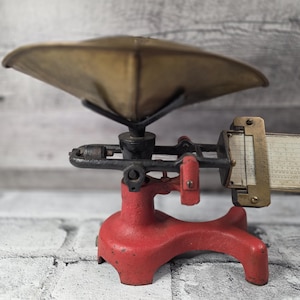 May include: Antique red and black scale with a brass pan. The scale has a vintage design with a sliding weight indicator. The scale is likely used for measuring weight, and it is a decorative item.