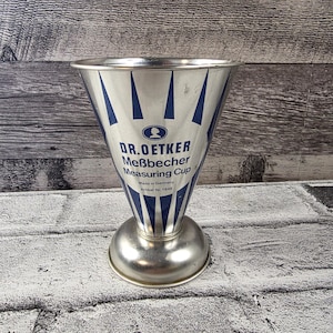 Peut inclure: Un ancien verre mesureur Dr. Oetker en métal argenté. Le verre a une forme conique avec des rayures verticales bleues et le texte "Dr. Oetker Meßbecher Measuring Cup". La base est un piédestal arrondi.