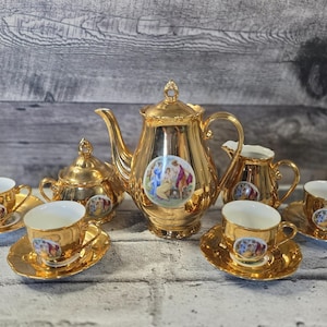 May include: A gold tea set comprising a teapot, sugar bowl, creamer, and four teacups with saucers. Each item features a painted scene. The set is displayed against a weathered wooden backdrop.