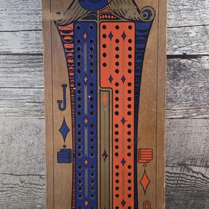 May include: Wooden cribbage board with a decorative design featuring blue and orange accents. The board has a metal hanger and is designed with playing card symbols. A classic game for family fun.