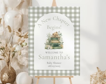 May include: A baby shower welcome sign on a wooden easel. The sign has a green and white gingham border. The center features the text "A New Chapter Begins!" above a watercolor illustration of a teddy bear on books. The sign reads "Welcome to Samantha's Baby Shower 08/05/2025."