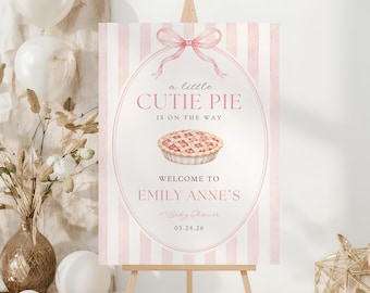 Op de afbeelding: Een rechthoekig bord met een roze en wit verticaal gestreepte achtergrond en een roze strikillustratie bovenaan. De tekst luidt: a little CUTIE PIE IS ON THE WAY WELCOME TO EMILY ANNE'S Baby Shower 05.24.26, met een taartafbeelding in het midden.