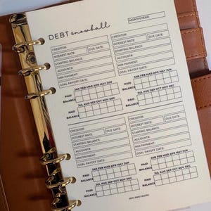 Op de afbeelding: Een plannerinzet met de woorden "DEBT snowball" bovenaan. De inzet heeft secties voor crediteuren, rentetarieven, vervaldatums en betalingsregistratie. De inzet zit in een bruine leren map met gouden ringen.