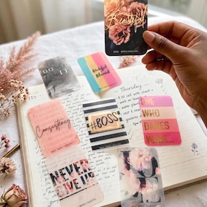 May include: A collection of rectangular cards with various designs and text including Beautiful, Don't Quit, Grateful Blessed, Consistency, #BOSS, She Who Dares Wins, Never Give Up, and Love, arranged on an open notebook with handwritten daily tasks.