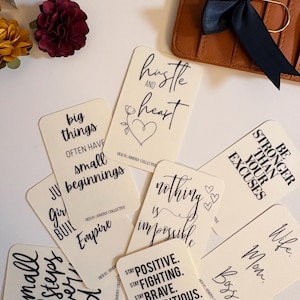 May include: Cream-coloured cards with black script displaying motivational quotes such as "Hustle and Heart" and "Nothing is Impossible." A brown leather wallet with a black ribbon is in the background. Decorative flowers are also present.