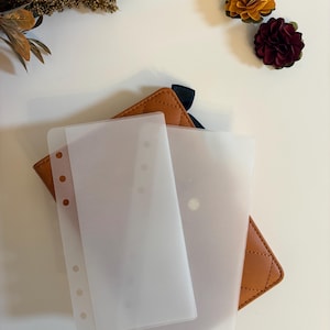 Könnte beinhalten: Klare Kunststoff-Ordner-Einsätze liegen auf einem braunen, gesteppten Leder-Notizbuch-Einband mit Rautenmuster. Dekorative gelbe und burgunderfarbene Blumen befinden sich im Hintergrund.