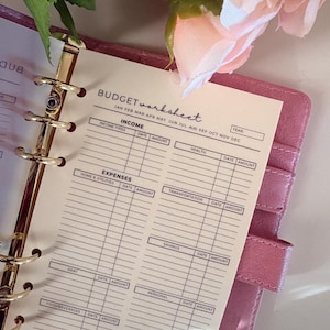 Peut inclure: Un planificateur budgétaire rose avec un classeur doré et une page "BUDGET worksheet". La feuille de calcul comprend des sections pour les revenus, les dépenses et l'épargne, avec des lignes pour les dates et les montants. Une rose rose et des feuilles vertes sont en arrière-plan.