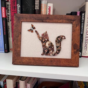 May include: A framed artwork featuring a squirrel made of buttons, reaching for a butterfly. The squirrel is brown and tan, with a fluffy tail. The butterfly is brown and orange. The frame is a dark brown color.