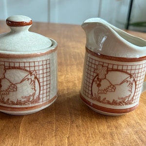 May include: A cream-colored ceramic sugar bowl with a lid and a creamer. Both pieces feature a brown grid pattern and a chicken illustration. The set is designed for serving sugar and cream.