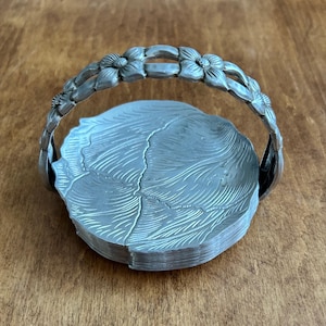 May include: A silver-toned metal basket with a floral handle and a leaf-like design on the base. The handle is adorned with flower details. The basket is likely used for serving or display.