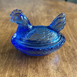 May include: A cobalt blue glass hen-on-nest dish. The hen is perched atop a basket-weave base, with detailed feather and comb textures. The lid is removable, revealing a small storage space. A decorative kitchen item.
