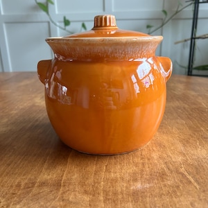 May include: A round, orange ceramic casserole dish with a lid. The dish has two small handles and a textured lid handle. The exterior is glossy, reflecting light. The dish is sitting on a wooden surface.