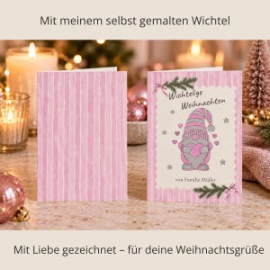 May include: A folded greeting card with a pink and white vertical striped pattern. The front features a gnome illustration, pink hearts, grey stars, pine branches, and the text Wichtelige Weihnachten von Familie Müller.