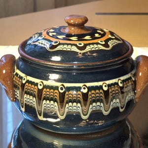 May include: A dark blue ceramic casserole dish with a lid. The dish has brown handles and a decorative pattern of white, brown, and orange wavy lines and circles. The lid also features a brown knob and similar patterns.