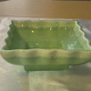 May include: A square, light green ceramic bowl with a scalloped rim and a rectangular base. The bowl has a glossy finish and a lighter, off-white color along the rim. The bowl is empty and sits on a white surface.